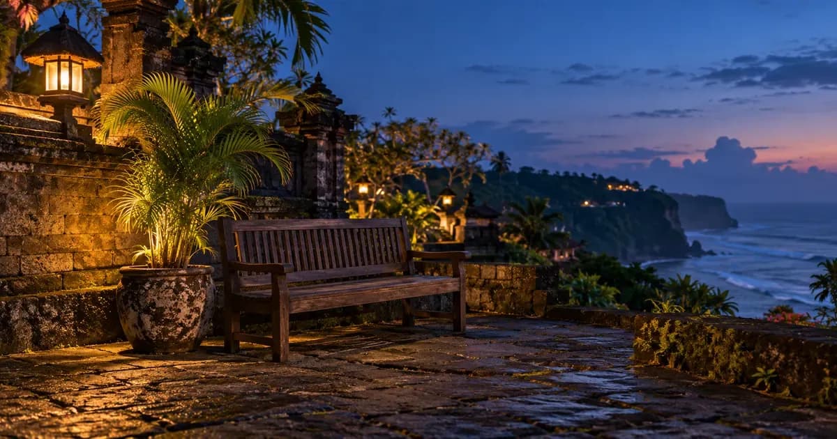 An empty waiting bench of dark timber beside a potted areca palm in Bali, weathered volcanic stone and moss in the foreground, at blue hour after sunset.