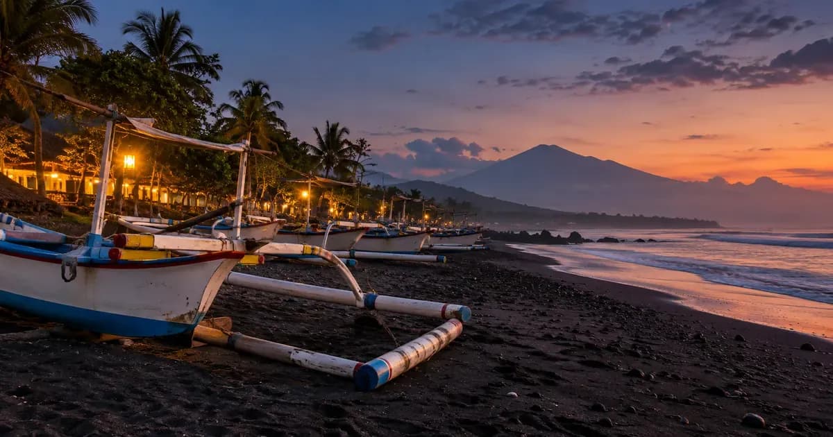 A black-sand beach with a line of outriggers drawn up above the tideline in Bali, warm haze softening the far distance, at blue hour after sunset.