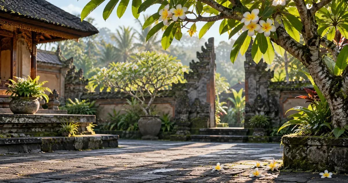 Close detail of a shaded courtyard of volcanic stone paving with a frangipani tree in Bali, overhanging frangipani branches framing one edge, in soft first light after dawn.