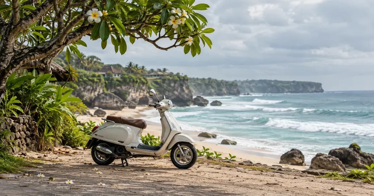 Wide view of a scooter parked on a sandy shoulder where a lane meets the beach in Bali, overhanging frangipani branches framing one edge, under a soft overcast sky.